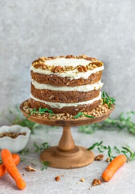 Side view of Keto Carrot Cake on wooden cake stand with chopped pecans and carrots