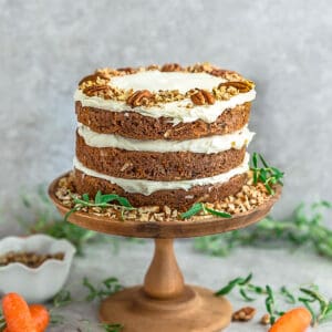 Side view of Keto Carrot Cake on wooden cake stand with chopped pecans and carrots