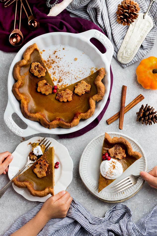 Two plates with a slice of pumpkin pie and a fork on each, with a hand holding one of the forks, next to three-quarters of a pumpkin pie in a pie dish