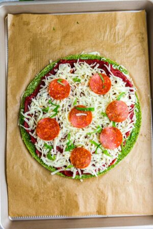 Top view of unbaked keto broccoli Pizza Crust with tomato sauce, cheese and pepperoni on brown parchment paper on a baking sheet