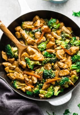 Close-up of stir fried cubed chicken with broccoli, carrots and a sesame sauce in a white pan with a wooden spoon