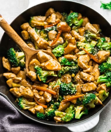 Close-up of stir fried cubed chicken with broccoli, carrots and a sesame sauce in a white pan with a wooden spoon