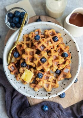 Top view of gluten-free waffle cereal in a white bowl with blueberries, butter, milk and maple syrup and a gold spoon