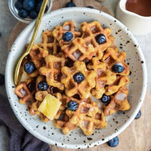 Top view of gluten-free waffle cereal in a white bowl with blueberries, butter, milk and maple syrup and a gold spoon