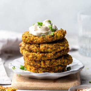 Side view of low carb zucchini fritters stacked on a white plate with a dollop of sour cream