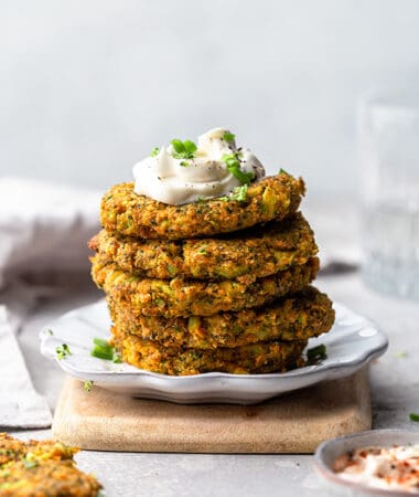 Side view of low carb zucchini fritters stacked on a white plate with a dollop of sour cream