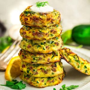 Side view of six keto zucchini fritters stacked on a white plate with lemon wedges