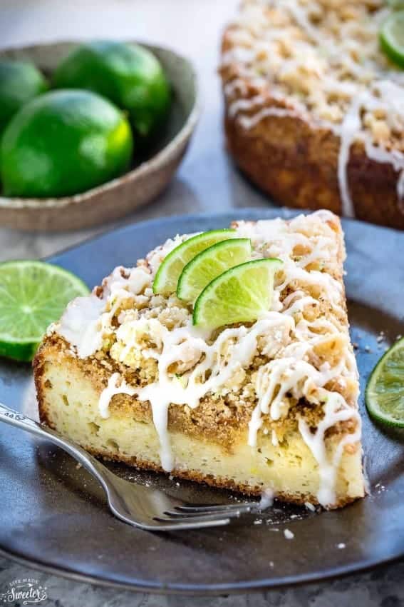 A slice of Key Lime Coffee Cake with streusel topping, white chocolate drizzle, and lime garnish on a plate with a fork