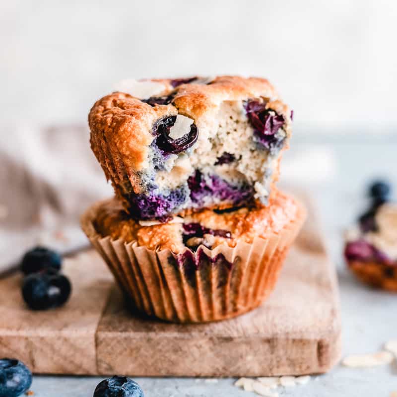 Side view of two half-eaten stacked Keto Blueberry Muffins on a wooden cutting board.