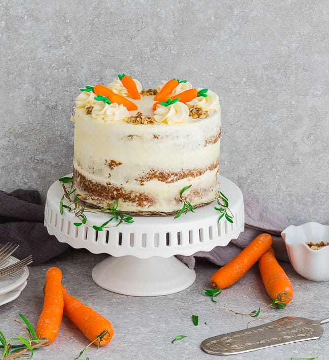 Side view of Keto Carrot Cake on wooden cake stand with chopped pecans and carrots