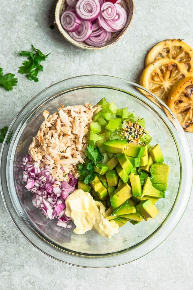Top view of ingredients to make chicken salad in a clear bowl on a grey background with lemon, oninos, celery, avocado, chicken and Whole30 mayo