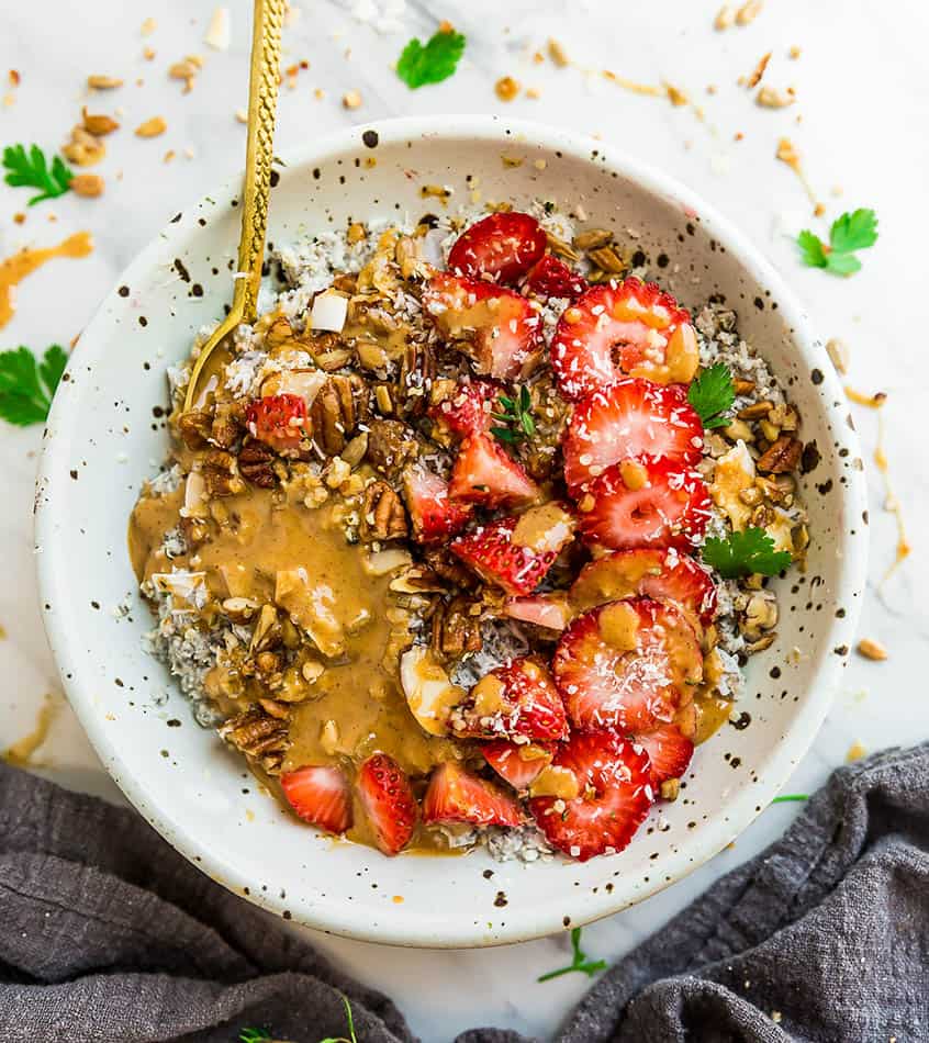 Top view of Low Carb Keto Overnight Oats in a white speckled bowl with strawberries, almond butter with a gold spoon on a white background