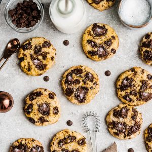 10 pumpkin chocolate chip cookies on a board with milk, a whisk and chocolate chips on the side