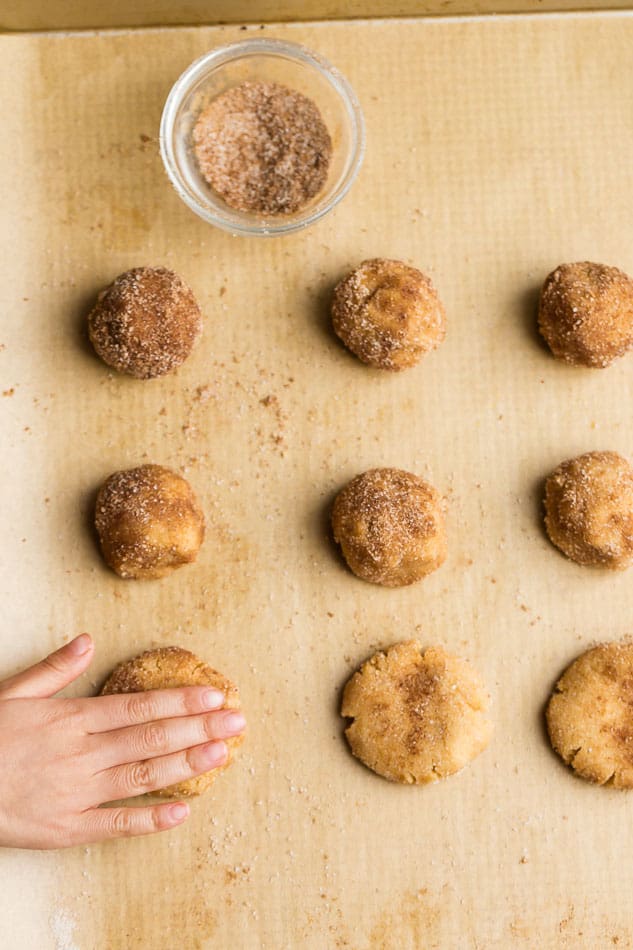 Top view of unbaked low carb snickerdoodle cookie doughballs with cinnamon sugar bowl on parchment paper on a baking sheet
