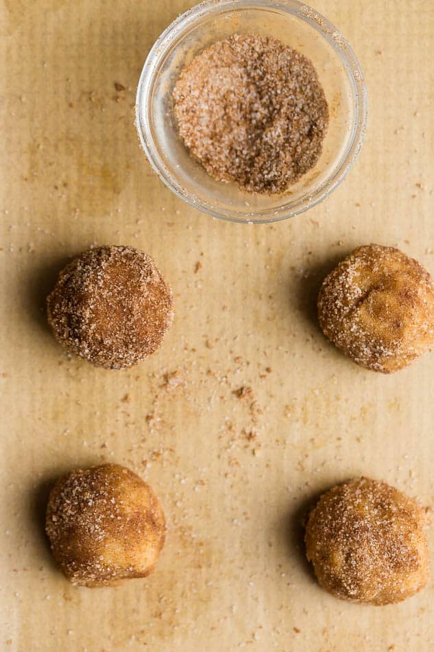 Four balls of snickerdoodle cookie dough, covered in cinnamon and sugar, on parchment paper, next to a bowl of cinnamon and sugar