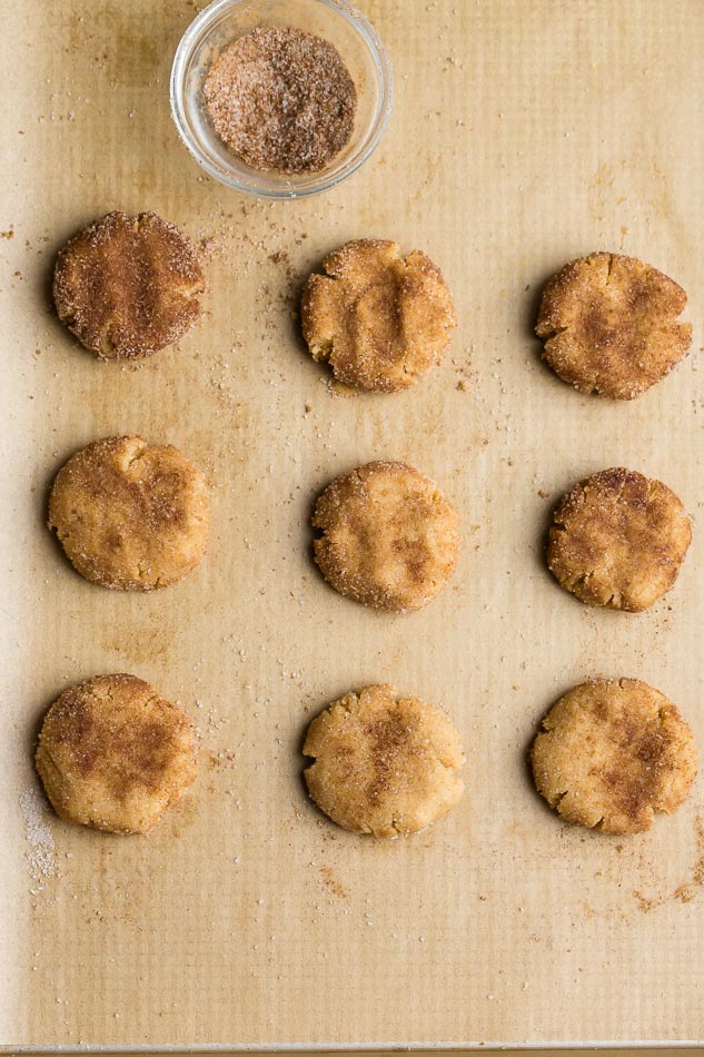 Overhead view of nine unbaked snickerdoodle cookies on parchment paper, coated in cinnamon and sugar, and pressed into flat disks, next to a bowl of cinnamon sugar 