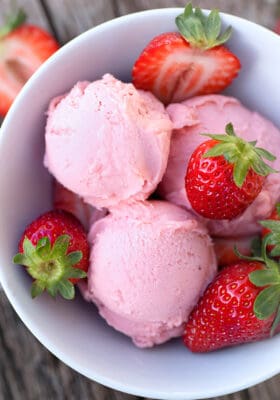 Top view of 3 scoops of low carb keto strawberry ice cream in a white bowl with fresh strawberries on a wooden background