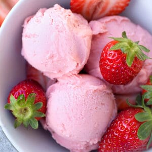 Top view of 3 scoops of low carb keto strawberry ice cream in a white bowl with fresh strawberries on a grey background
