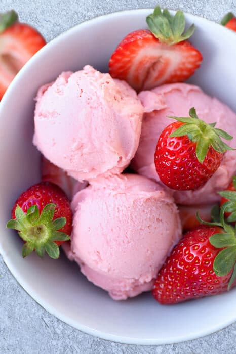 Top view of 3 scoops of low carb keto strawberry ice cream in a white bowl with fresh strawberries on a grey background