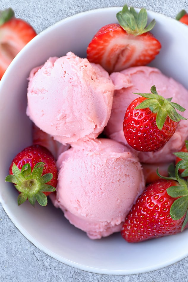 Top view of 3 scoops of low carb keto strawberry ice cream in a white bowl with fresh strawberries on a grey background for berry recipes.