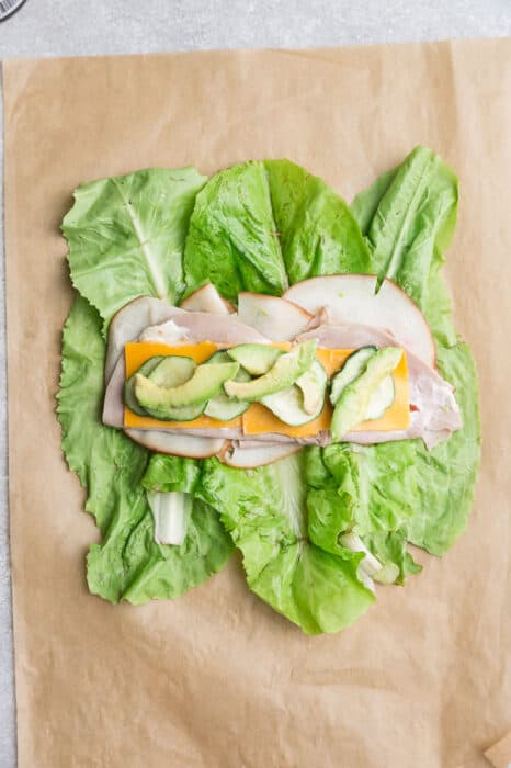 Top view of lettuce layered with turkey, ham, avocado, cheese, cucumbers and mayonnaise on a brown parchment paper.