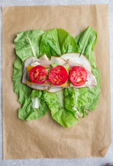 Top view of lettuce layered with turkey, ham, tomatoes, cheese, cucumbers, avocado, mayonnaise on a brown parchment paper.