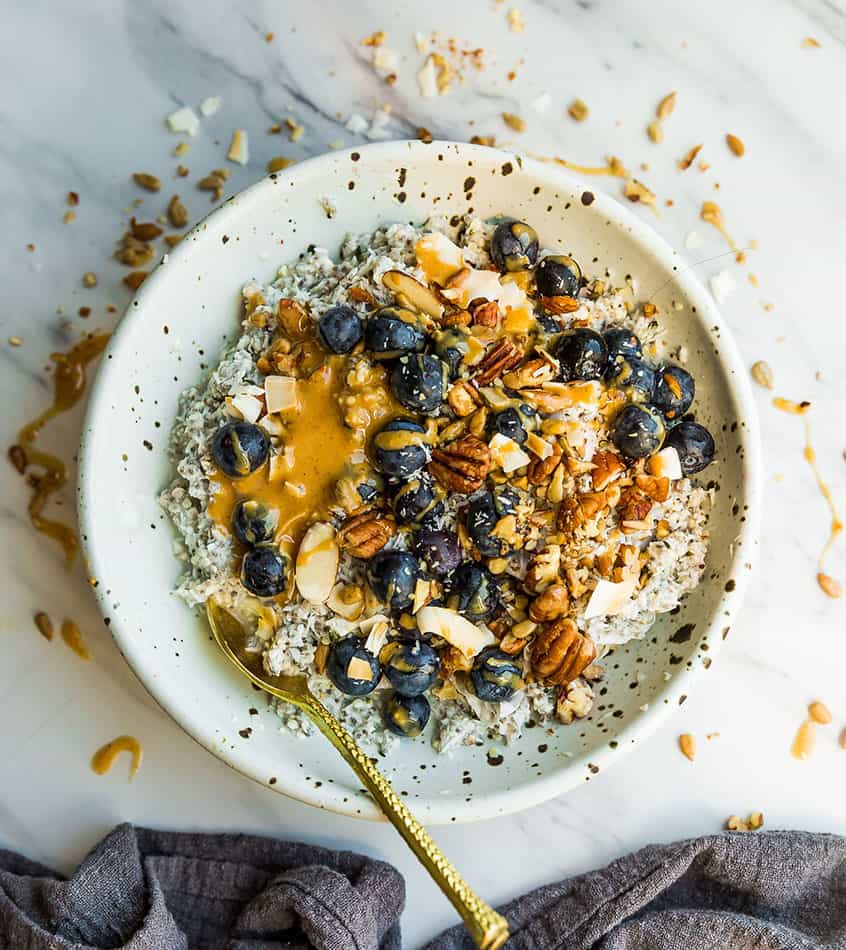 Top view of Low Carb Keto Overnight Oats in a white speckled bowl with blueberries, almond butter with a gold spoon on a white background
