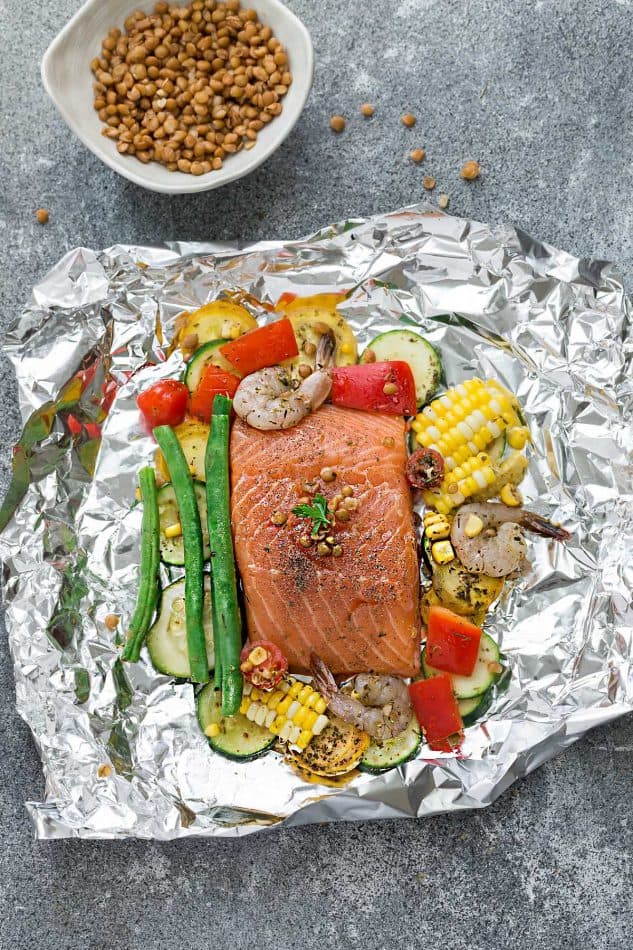 Top view of uncooked ingredients for Mediterranean Salmon Foil Packets on a sheet of foil next to a bowl of lentils