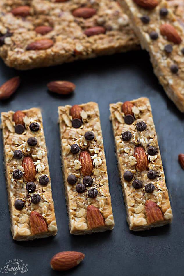 Three granola bars in a row, topped with almonds and chocolate chips, with other granola bars in the background.