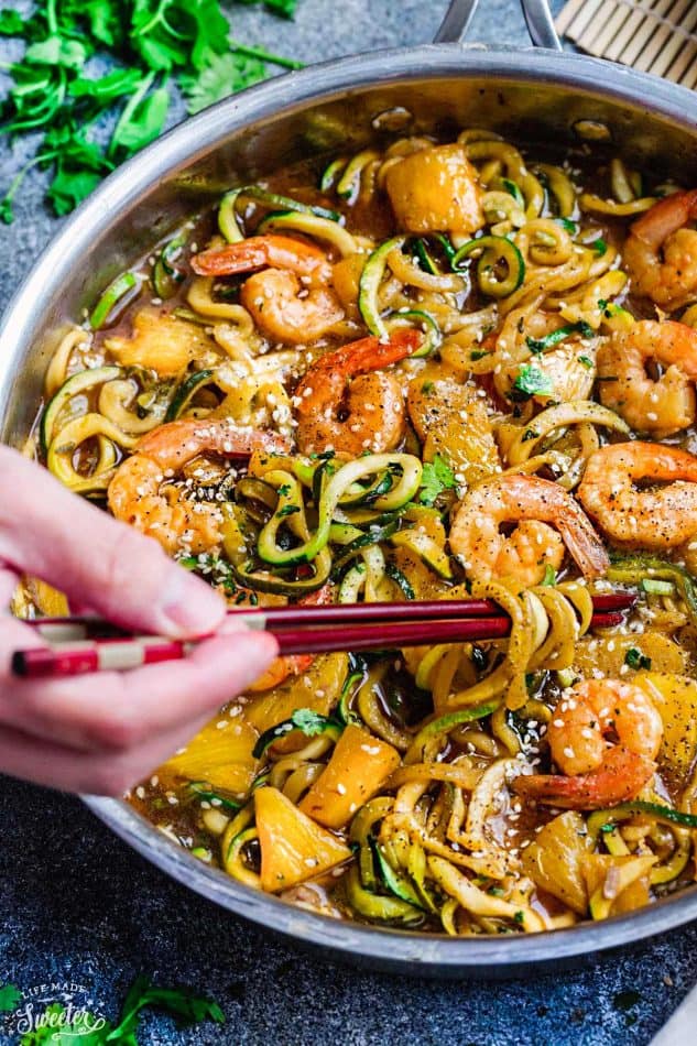 Close-up view of teriyaki shrimp with zucchini noodles in a stainless steel pan with a hand holding chopsticks