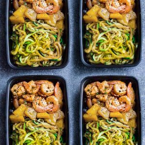 Top view of healthy shrimp teriyaki with zucchini noodles in four black meal prep containers on a grey background