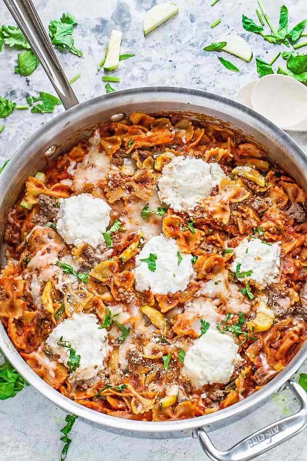 A stainless steel pot of skillet lasagna topped with ricotta and parsley, next to spinach leaves, parsley leaves, zucchini slices, and a wooden spoon 