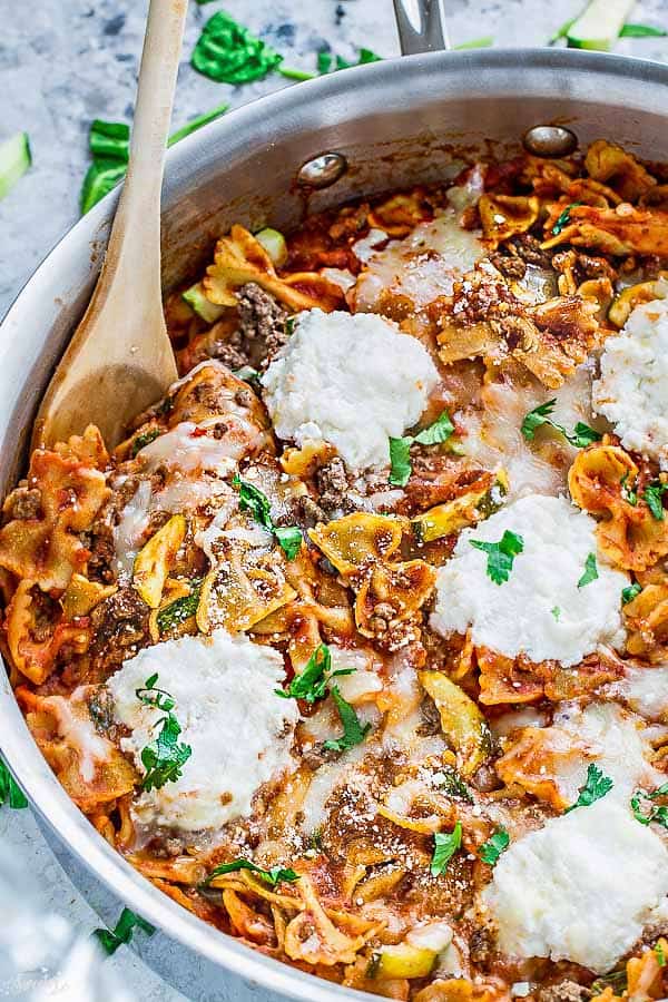 Close up of a pot of skillet lasagna topped with ricotta and chopped parsley, with a wooden spoon in it.