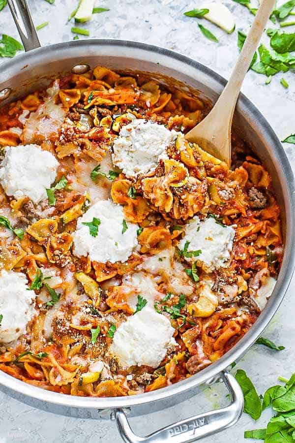 A pot of skillet lasagna topped with ricotta cheese with a wooden spoon in the bowl, next to leaves of spinach.