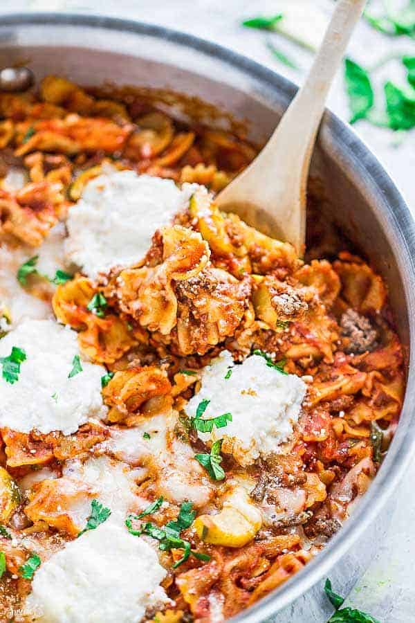 Close up of a wooden spoon in a pot of skillet lasagna, topped with ricotta and parsley, with basil leaves in the background. 