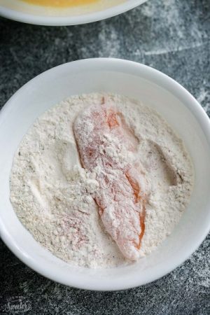 A piece of raw chicken being coated in flour mixture in a bowl