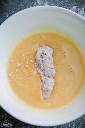 A piece of flour-coated raw chicken being dredged in a bowl of egg mixture