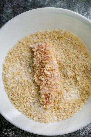 A piece of raw chicken being coated in a bowl of crispy breading