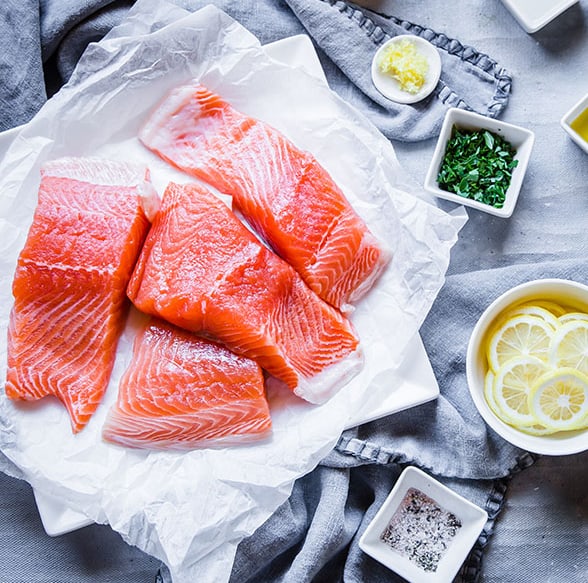 Raw salmon fillets on a plate lined with parchment paper beside a bowl of fresh lemon slices