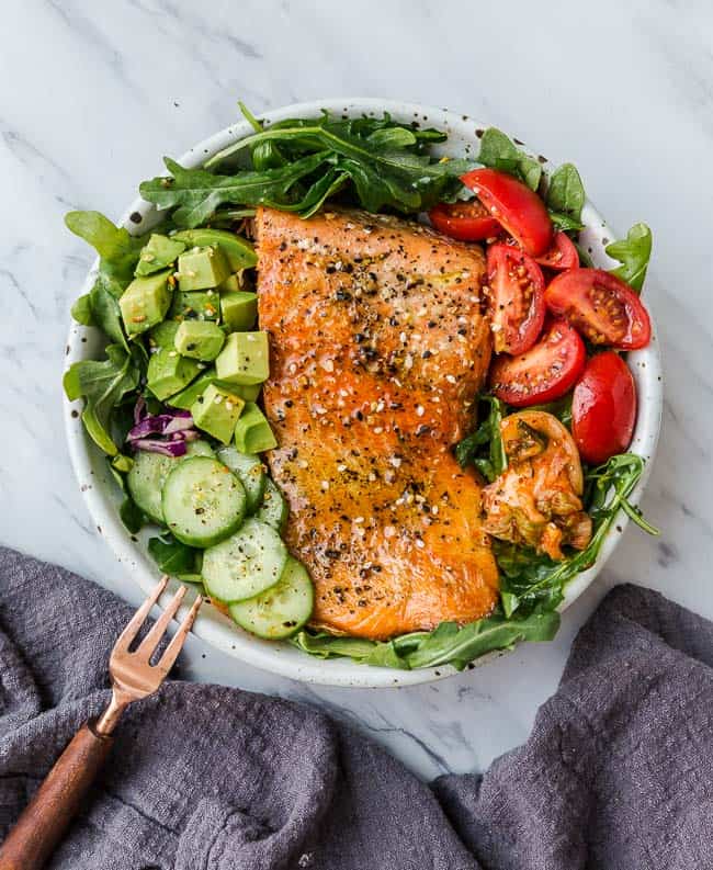 A bowl of baked salmon surrounded by tomatoes, cucumbers and avocado slices