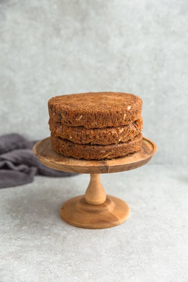 Three cooled layers of carrot cake stacked on top of a wooden cake stand