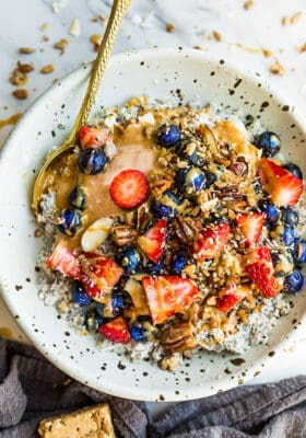 Top view of Low Carb Keto Overnight Oats in a white speckled bowl with berries, almond butter with a gold spoon on a white background