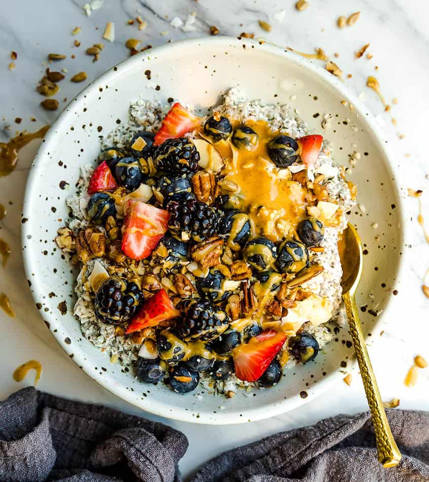 Top view of Low Carb Keto Overnight Oats in a white speckled bowl with berries, almond butter with a gold spoon on a white background