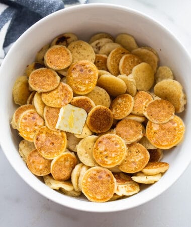 Top view of mini pancake cereal in a white bowl with a square of butter and maple syrup