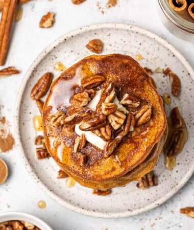 Overhead view of a stack of pumpkin pancakes topped with pecans, butter and maple syrup