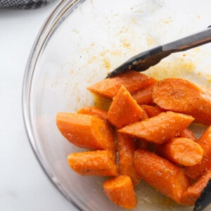 Overhead view of sliced raw carrots in a clear mixing bowl tossed with salt, black pepper, oil and coconut aminos with tongs.