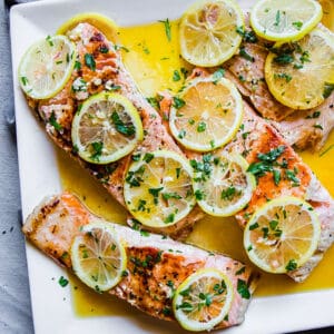 A close-up shot of pan-fried fish fillets on a white plate with fresh parsley and lemon slices on top