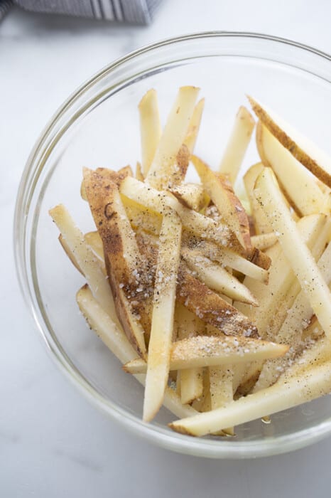 Potatoes cut into fries with seasoning in a clear bowl