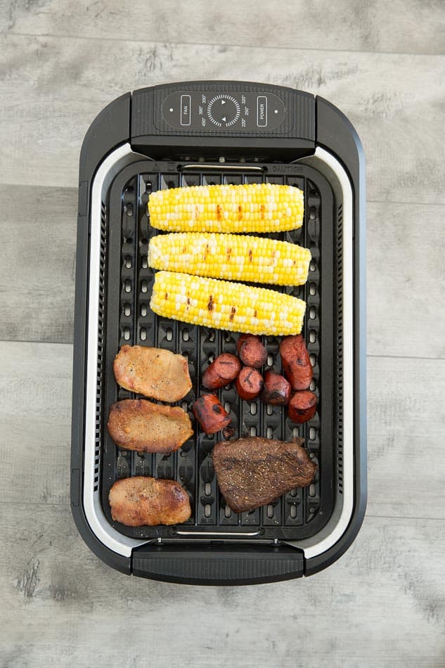 Overhead view of pork chops, steak, chunks of sausage and ears of corn in a Power Smokeless Grill