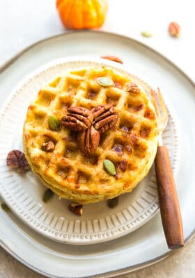 Top view of 3 pumpkin chaffles on a white plate with pecans, pumpkin seeds and sugar free syrup on a white plate with a fork